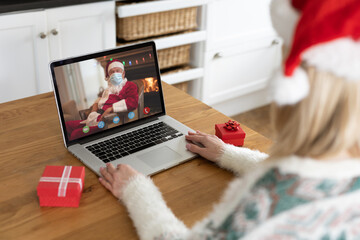 Wall Mural - Caucasian woman making laptop christmas video call with santa claus in face mask