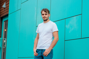 A young man in a white blank T-shirt stands against the background of a blue wall.