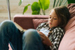© dikushin - Side view of tired adorable child kid girl reading paper book lying on soft couch at home looking away. Portrait of little cute upset girl lying on sofa alone in cozy living room.