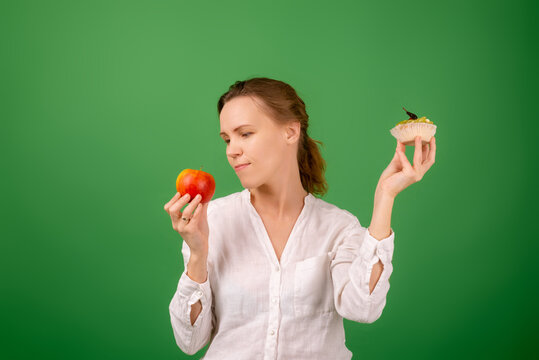 Middle-aged woman with apple and cake on a green background. Looks at the apple. The concept of choosing the right healthy food.