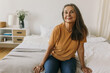© shurkin_son - Indoor image of beautiful senior female in casual yellow T-shirt and blue jeans sitting on white bed in her bedroom smiling, having rest at home during weekend, waiting grandchildren to visit her
