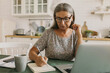 © shurkin_son - Charming mature woman in stylish glasses and romantic-looking dress sitting in kitchen, studying online, noting down information while watching English lessons on her laptop. Education and technology