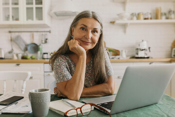 Adorable charming elderly female in dress holding hand over cheek smiling warmly at camera while resting from work online on her laptop, phone with copy space for your advertisement lying next to her