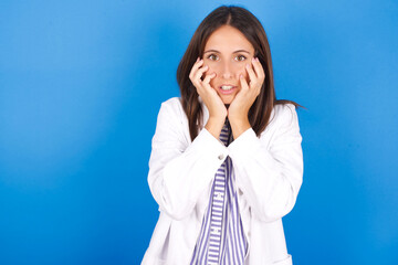 Wall Mural - Stupefied young european doctor girl wearing white medical gown ​on white background expresses excitement and thrill, keeps jaw dropped, hands on cheeks, has eyes popped out