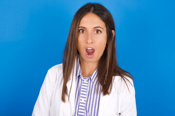 Wall Mural - Oh my God. Surprised young european doctor girl wearing white medical gown ​on white background stares at camera with shocked expression exclaims with unexpectedness,