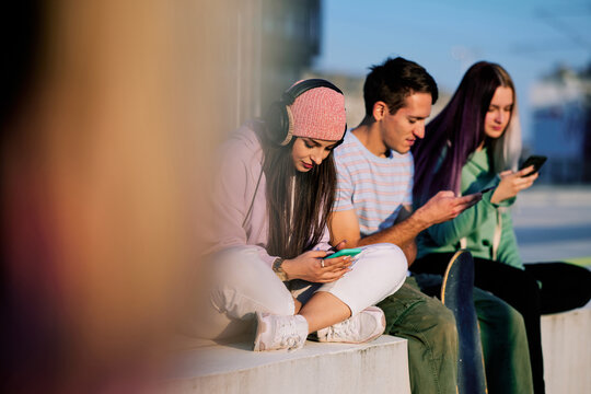 Three teenage friends are sitting in the urban exterior and hanging on their phones.