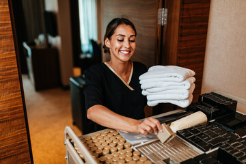 Beautiful young hotel chambermaid in uniform bringing clean towels and other supplies to hotel room.