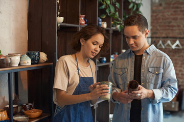 Poster - Positive young saleswoman in apron standing against shelves and showing ceramic mug to buyer in pottery store