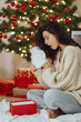 © Buyanskyy Production - Curious happy romantic woman sitting in front of Christmas tree background. Shocked lady surprised of the present after the opening in the gift box