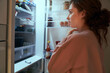 © gpointstudio - Undecided young caucasian woman checking fridge for some food at night