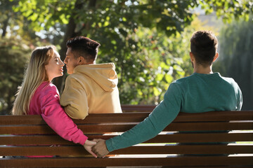 Wall Mural - Woman holding hands with another man behind her boyfriend's back on bench in park. Love triangle