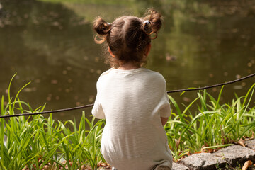 Little cute redhead girl with ponytails sits on the shore of a pond with her back to the camera