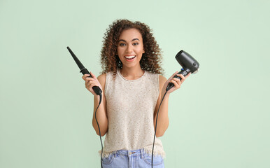 Happy young African-American woman with curling iron and hair dryer on green background