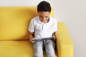 Poster - Little African-American boy with tablet computer watching cartoons on armchair at home