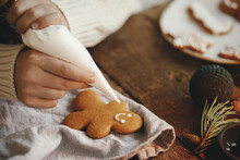 Hands Decorating Christmas Gingerbread Man Cookie With Frosting On Rustic Table With Napkin, Spices, Decorations. Atmospheric Moody Time. Making Traditional Christmas Gingerbread Cookies
