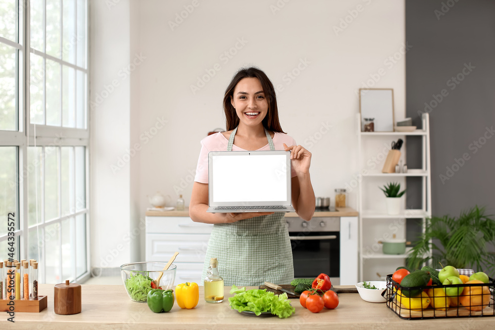 Young woman with fresh vegetables and laptop in kitchen