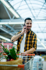 Wall Mural - Young handsome man in a supermarket using smartphone while grocery shopping