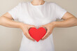Cropped closeup photo of girl in white t-shirt holding red paper heart near upper abdomen on isolated grey background