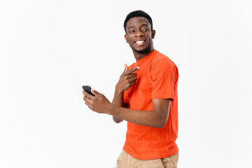 young man of african appearance with a mobile phone and in an orange t-shirt on a light background c