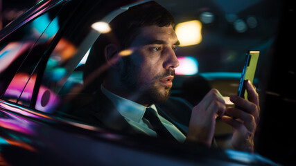 Wall Mural - Handsome Businessman in a Suit Commuting from Office in a Backseat of His Luxury Car at Night. Entrepreneur Using Smartphone while in Transfer Taxi in Urban City Street with Working Neon Signs.