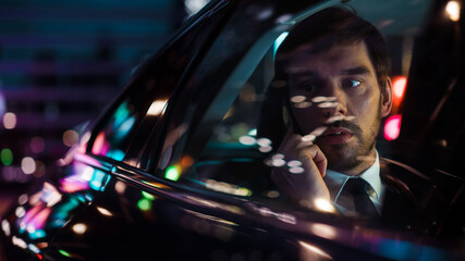 Wall Mural - Handsome Businessman in a Suit Commuting from Office in a Backseat of His Car at Night. Entrepreneur Talking on Smartphone while in Taxi in Urban City Street with Working Neon Signs.