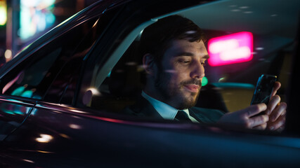 Wall Mural - Handsome Businessman in a Suit Commuting from Office in a Backseat of His Luxury Car at Night. Entrepreneur Using Smartphone while in Transfer Taxi in Urban City Street with Working Neon Signs.