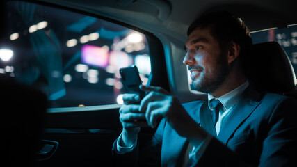 Successful Businessman in a Suit Commuting from Office in a Backseat of His Luxury Car at Night. Entrepreneur Using Smartphone while in Transfer Taxi in Urban City Street with Working Neon Signs.