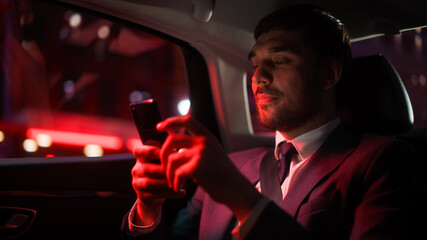 Wall Mural - Successful Businessman in a Suit Commuting from Office in a Backseat of His Car at Night. Entrepreneur Using Smartphone while in Transfer Taxi in Urban City Street with Working Neon Signs.