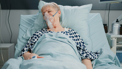 Retired woman with oxygen tube against respiratory problem in hospital ward bed. Sick patient breathing heavily while resting and waiting for medical assistance with IV drip bag.