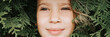 © Ksenia - portrait of the face of a cute happy caucasian candid healthy eight year old kid girl surrounded by branches and leaves of green plant thuja or cypress on nature outdoor. banner