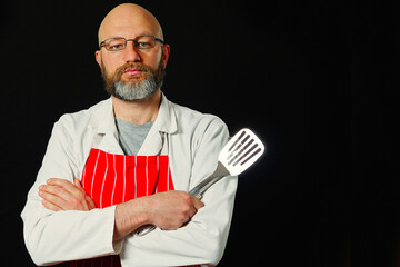 Portrait of a bald cook in his 40s on dark background. Man in white uniform and red and white stripe classic apron holding flipper and ladle, wearing glasses. Slim body type, friendly face expression