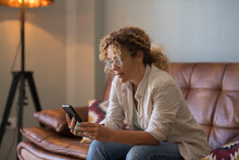 Serious Young Woman Text Messaging Using Mobile Phone While Sitting On Sofa At Home. Woman With Curly Hair And Eyeglasses Reading Or Texting Using Mobile Phone In The Living Room