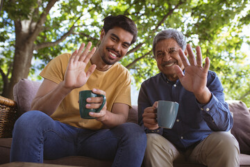 Happy biracial adult son and senior father holding cups of coffee and making laptop video call