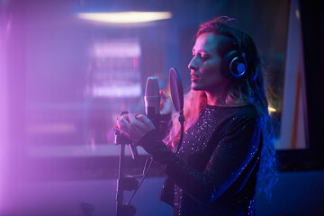 Young singer listening to music in headphones and singing a song to microphone at the recording studio