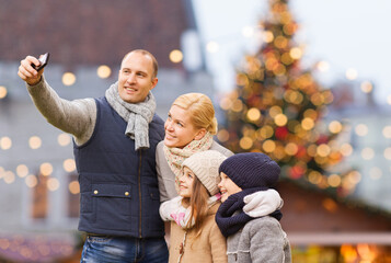 Wall Mural - winter holidays, technology and people concept - happy family taking selfie with smartphone over christmas market background