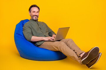 Sticker - Portrait of attractive cheery man sitting in bag chair using laptop typing project isolated over bright yellow color background