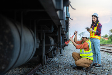 Man engineer railway wearing safety uniform and helmet using a wrench to repair and inspect the undercarriage of the train. Women engineer holding tablet for inspection.