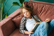 High-angle view of sad adorable child kid girl reading paper book lying on soft couch at home looking away. Top view of little cute upset girl lying on sofa alone in cozy living room.