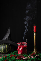 View of red halloween cocktail in glass with straw with lit candle and pumpkin with witch hat, on decorated table, black background, vertical