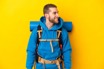 Young caucasian mountaineer man with a big backpack isolated on yellow background looking side