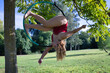 © Manuel - Young blonde woman in a red party dress exercising on an aerial hoop hanging from a tree in the middle of a park. Concept aerial hoop, pole dance, fitness, curvy girl.