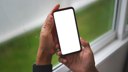 Poster - Photo of a woman hand with a digital watch holding a white blank screen smartphone over the window sill as a background.