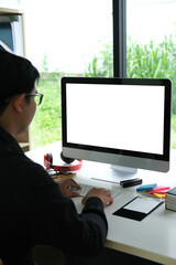 Poster - Portrait of young programmer typing on a keyboard at the white table surrounded by a blank screen computer monitor, sticky notes, headphones, glasses, clipboard, book and stapler.