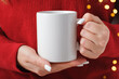 © Marykor - Female hands hold a mockup white tea mug in a red winter sweater, gold bokeh