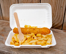 Close And Selective Focus On A Polystyrene Carton Comprising A Battered Jumbo Sausage And Traditional Chip Shop Chips Cooked In Beef Dripping On An Outdoor Wooden Table
