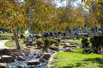 two women with baby stroller sitting on lush green grass in the park surrounded by autumn trees near a running river at Kenneth Hahn Recreation Area in Los Angeles California USA