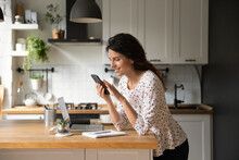 Happy Latin freelance employee working from home, using online app on smartphone at laptop in kitchen, smiling at video call, texting on social media chats, reading article. Digital life concept