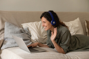 Wall Mural - Happy woman in headphones with laptop making video call, waving hello, speaking and smiling at screen, while resting on couch at home during distance conference meeting with family, relatives, friends