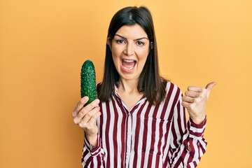 Young hispanic woman holding cucumber pointing thumb up to the side smiling happy with open mouth