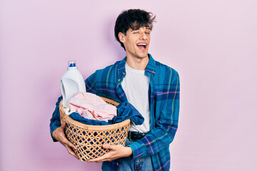 Poster - Handsome hipster young man holding laundry basket and detergent bottle winking looking at the camera with sexy expression, cheerful and happy face.